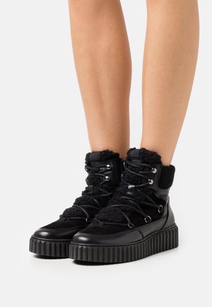 Schwarze High-Top-Plateau-Stiefel mit dicken, gerippten Sohlen, Schnürfronten und strukturierten Stoffdetails, getragen an nackten Beinen.