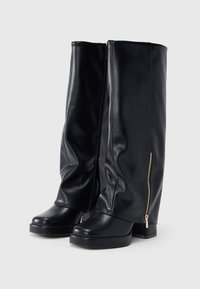 Bottes plates-formes hautes en cuir noir avec talons carrés et fermetures éclair latérales, présentées sur un fond clair uni.