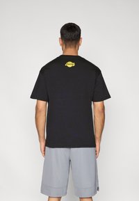 Μαύρο βαμβακερό μπλουζάκι με στρογγυλή λαιμόκοψη και κοντά μανίκια, με κίτρινο τυπωμένο λογότυπο στο επάνω μέρος της πλάτης. Φοράει με γκρι σορτς.