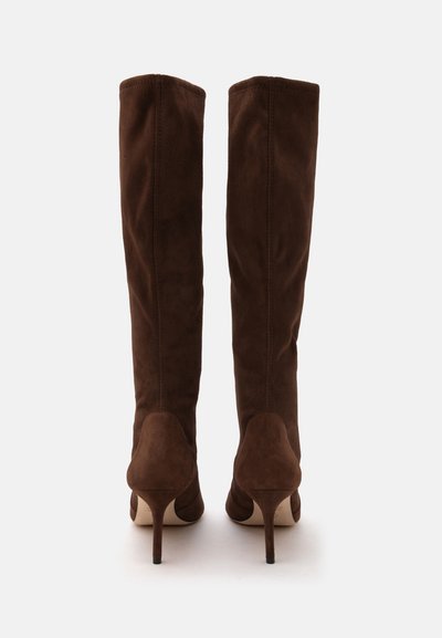 Paire de bottes hautes en daim marron à talons, vues de dos sur un fond clair uni.