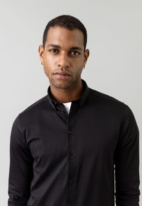 Melna garākā piedurkņu krekls ar pogājamu apkakli, kas izceļas ar gludu tekstūru un pieguļošu dizainu, akcentēts ar melnām pogām.