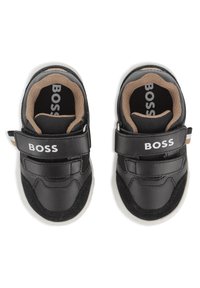 Chaussures en simili cuir noir avec doublure en tissu beige, dotées de brides auto-agrippantes et de semelles en caoutchouc blanc. Marque "BOSS" visible sur les brides et les semelles intérieures.