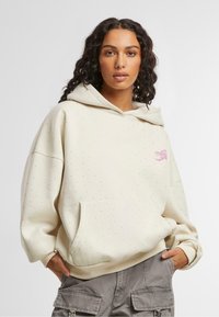 Donna con lunghi capelli ricci che indossa un hoodie beige chiaro con piccoli puntini e pantaloni cargo grigi, con le mani nelle tasche.