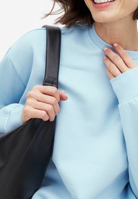 Sudadera azul claro hecha de una tela suave; bolso de cuero negro sostenido con una mano desnuda. Detalles de puños y cuello acanalados visibles.