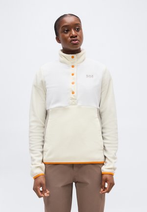 Naine, kes kannab kreemjat fliisist pusa oranžide nööpide ja äärisega, seisab valge lihtsa tausta ees, vaadates küljele.