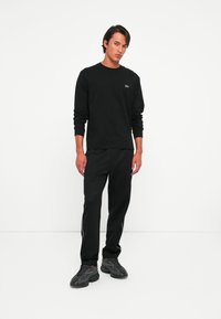 Man staat in een zwart langemouwen shirt met een klein logo, zwarte broek met witte zijkanten, en zwarte sport sneakers tegen een witte achtergrond.