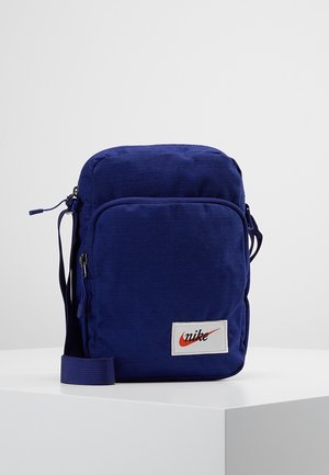 Petit sac bandoulière bleu compact Nike avec poche zippée avant et sangle réglable sur une surface blanche avec un fond uni.