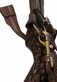 Close-up van een bruin gevlochten leren tasriem met een messing draaiklink en een ritsv pull-tab zichtbaar.