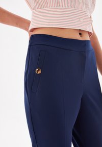 Pantaloni blu navy realizzati in un tessuto liscio, con una tasca laterale caratterizzata da un dettaglio in bottone dorato e una vestibilità su misura, che mostrano un design moderno.