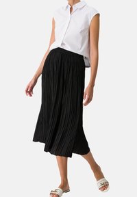 Jupe mi-longue plissée noire avec ceinture élastique, associée à un haut sans manches blanc à col et des sandales légères. Texture lisse sur toute la surface.