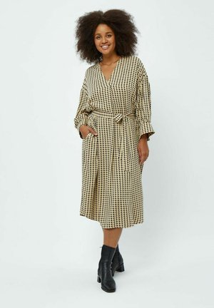 Robe beige avec un motif à carreaux noirs, décolleté en V, manches longues, taille cintrée, poches latérales et longueur midi, associée à des bottines noires.