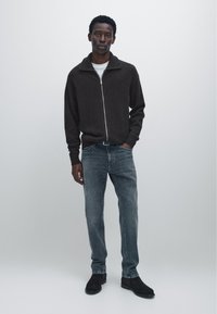 Donkerbruine zip-up sweater met hoge kraag, grijze spijkerbroek en zwarte suède laarzen. Model staat tegen een lichtgrijze achtergrond.