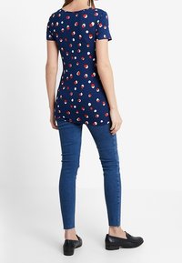 Vrouw in een marineblauwe short-sleeve shirt met rode en witte stippen, gecombineerd met strakke blauwe jeans en zwarte loafers, staand met de rug naar de kijker gekeerd.