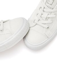 White canvas sneakers with textured rubber soles, round toe, and six eyelets for laces. Minimalist design with a clean, uniform color.