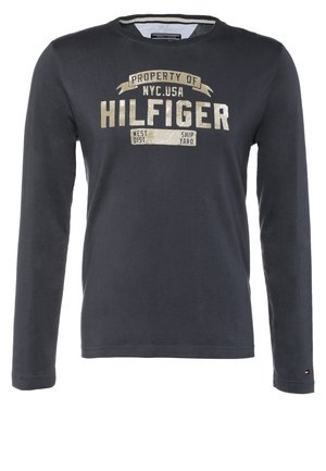 Man draagt marineblauw shirt met lange mouwen en een bruine broek, en draagt een grote beige reistas met bruine handvatten terwijl hij tegen een effen achtergrond loopt