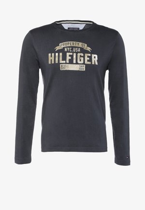 Man draagt marineblauw shirt met lange mouwen en een bruine broek, en draagt een grote beige reistas met bruine handvatten terwijl hij tegen een effen achtergrond loopt