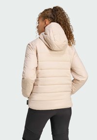 Eine Person mit lockigem Haar ist von hinten zu sehen, trägt eine hellbeige Daunenjacke mit Kapuze und schwarze Hosen vor einem hellen Hintergrund.