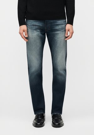 Moški v izpiranih modrih kavbojkah, črni majici z dolgimi rokavi in črnih usnjenih čevljih stoji ob preprostem ozadju.