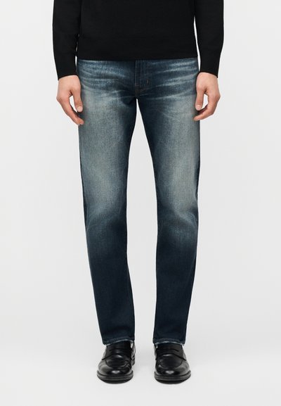 Homme portant un jean bleu délavé, un top noir à manches longues et des chaussures en cuir noir, debout devant un fond uni.