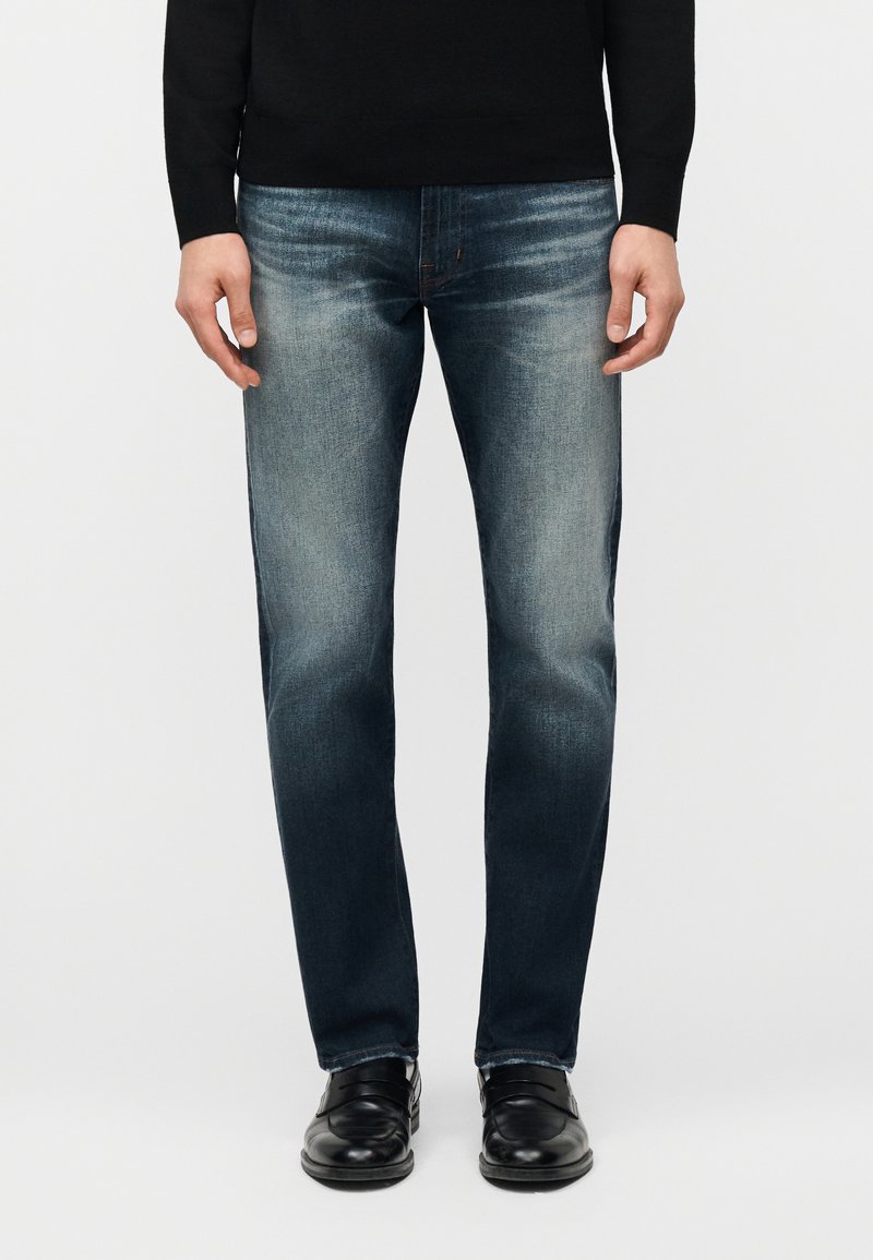 Homme portant un jean bleu délavé, un top noir à manches longues et des chaussures en cuir noir, debout devant un fond uni.