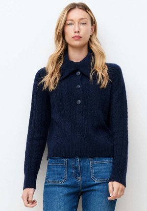 Kobieta o długich blond włosach, ubrana w granatowy sweter na guziki i niebieskie dżinsy, stojąca na tle jednolitego białego tła.