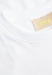 Bijela pamučna majica s okruglim izrezom koja ima glatku teksturu i oznaku s "JJXX" u žutoj boji na bež pozadini.