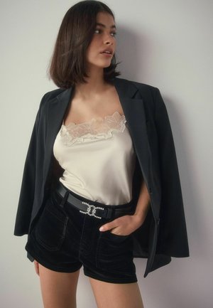 Schwarzer Blazer über einem cremefarbenen Satin-Cami mit Spitze, kombiniert mit schwarzen Samt-Shorts. Der Gürtel hat einen Perlenakzent. Glatte Texturen und gedämpfte Farben.