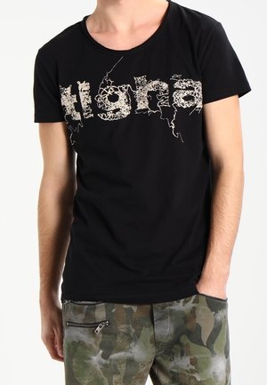 Mand iført en sort t-shirt med krøllet hvid tekst og camouflagebukser med en lynlåslomme på højre lår.