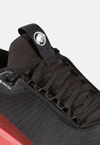 Czarne buty sportowe o teksturowanej cholewce, z czerwonymi akcentami i metką GORE-TEX. Krzyżowane sznurówki i detal z pętelką zwiększają funkcjonalność.