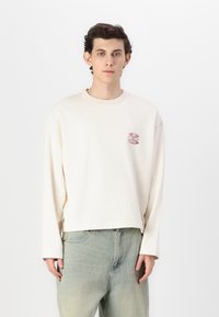 Camisa de manga larga beige claro con cuello redondo y un pequeño detalle gráfico rojo en el pecho, combinada con jeans azul claro.