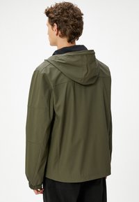 Ung mand med krøllet hår iført en olivengrøn hættejakke set bagfra mod en ensfarvet hvid baggrund.