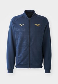 Veste zippée marine avec col montant, présentant un motif géométrique subtil et des accents de logo dorés sur la poitrine et la manche. Tissu doux.