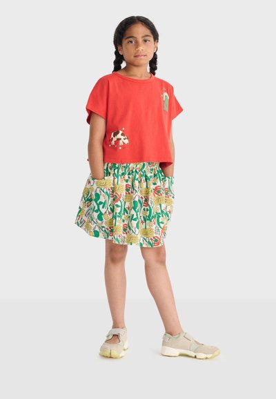 Fille aux cheveux tressés portant un chemisier rouge court avec des imprimés d'animaux de la ferme et une jupe fleurie à poches, debout les mains dans les poches.