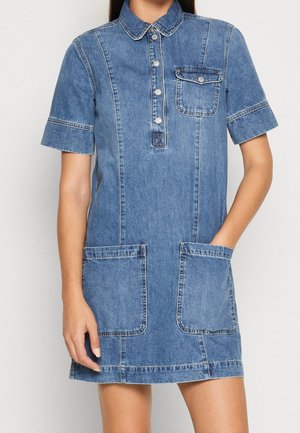 Kobieta w krótkim, jeansowym sukienka z kołnierzykiem, zapięciem na guziki z przodu, kieszonką na piersi oraz dwoma dużymi naszywanymi kieszeniami z przodu.