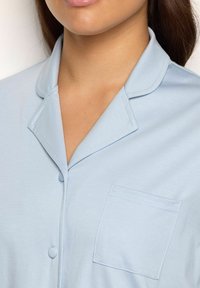 Camicia da pigiama azzurro chiaro con colletto, chiusura frontale con bottoni e taschino a toppa. Tessuto morbido con una trama liscia e dettagli di cucitura curati.