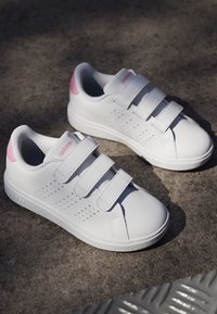 Zapatillas blancas con tres cintas de velcro, parte superior sintética suave, acentos rosas en el talón y patrón perforado para transpirabilidad.