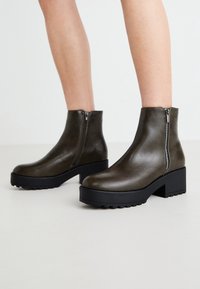 Botines de cuero verde oliva con puntera redonda, cremalleras laterales y suela de plataforma negra gruesa. Acabado suave con un tacón bajo y cuadrado.