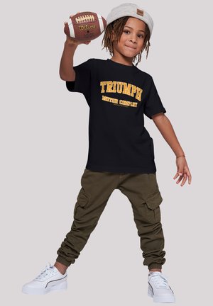 Schwarzes T-Shirt mit dem Grafik "TRIUMPH MOTOR COMPANY" in Gelb, olivgrüne Cargo-Hose, weiße Sneaker und graue Mütze. Kind hält einen American Football.