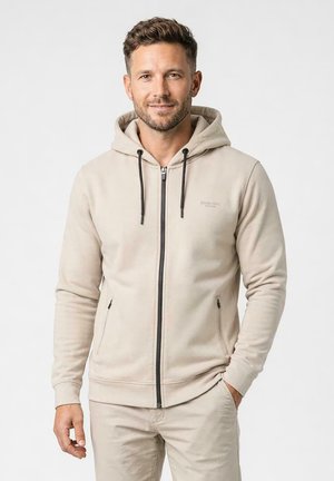 Man met kort krullend haar en baard, gekleed in een beige hoodie met rits en bijpassende broek, staand met één hand in de zak tegen een effen achtergrond.