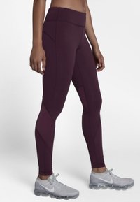 Nogi w ciemnofioletowych legginsach sportowych z wysokim stanem i jasnoszarych butach do biegania Nike, stojące na jednolitym tle.