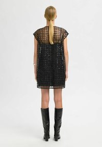 Moteris su ilgu šviesiaplaukiu uodegėle, vilkinti trumpą juodą blizgučių suknelę su permatoma tinklo danga ir juodais ilgaauliais batais, stovi nusisukusi.