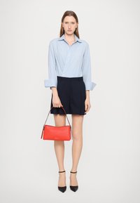 Chemise à rayures bleues et blanches, short taille haute bleu marine, escarpins noirs pointus et un sac à main rouge texturé avec une courte bandoulière.