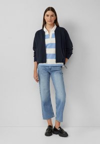 Junge Frau mit einer marineblauen Bomberjacke, einem blau-weiß gestreiften Polo, hellblauen, taillierten Jeans und schwarzen Slippern, die gegen eine graue Wand steht.