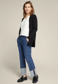 Cardigan aperto nero, top bianco a V, jeans blu con risvolti in denim. Il modello indossa mocassini neri. Look casual, vestibilità rilassata con tessuti a contrasto.