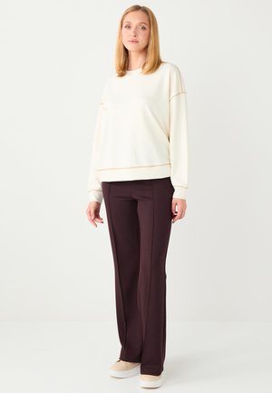 Giovane donna con capelli biondi lisci che indossa un maglione crema largo, pantaloni marrone scuro a gamba larga e scarpe da ginnastica beige, in piedi su sfondo bianco.