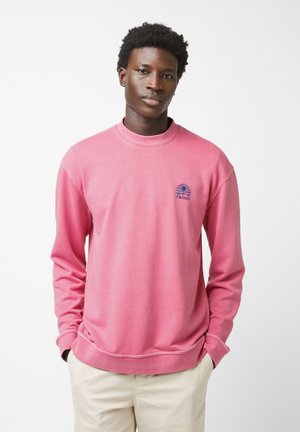 Felpa rosa con colletto rotondo e maniche lunghe. Presenta un logo blu navy sul petto. Realizzata in tessuto morbido con una texture liscia.