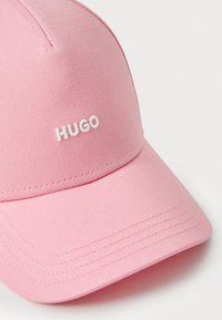 Pink cap lavet af tekstureret stof, med en buet skygge og hvid "HUGO" logo foran. Syninger synlige langs capens kanter.