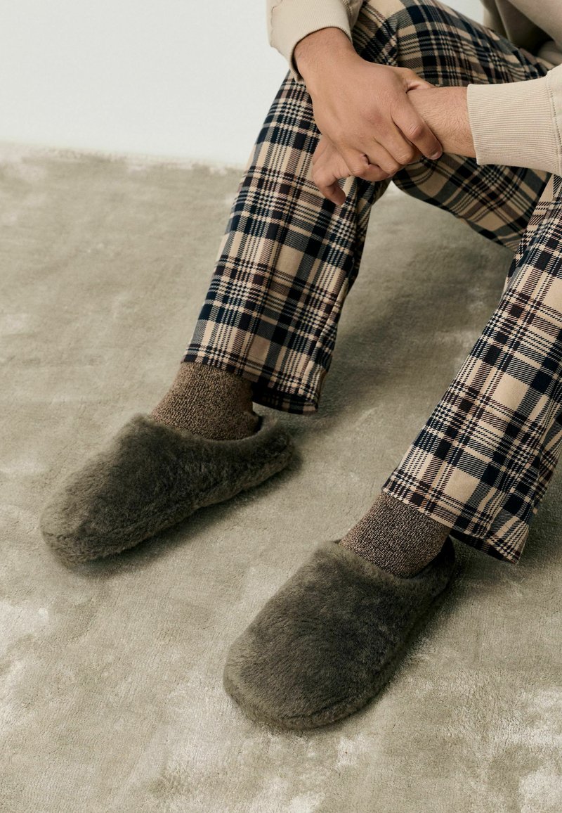 Grå, fluffiga tofflor med rundad tådesign, strukturerad yta och mjukt foder. Kombinerat med bruna mönstrade strumpor och rutiga byxor.