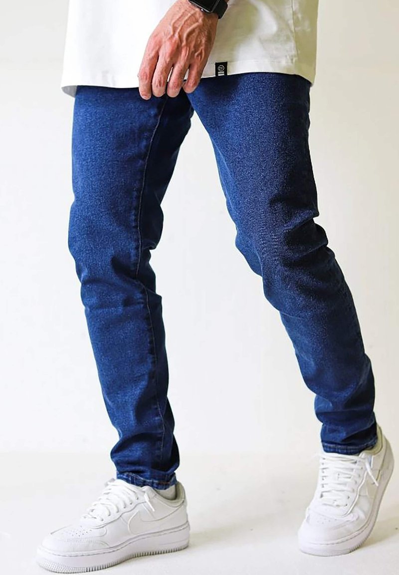 Personne portant un jean bleu slim, des baskets blanches et une chemise longue blanche, debout devant un fond clair uni.