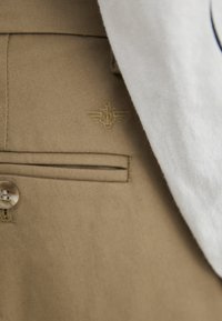 Tan trousers with a subtle stitched pocket, featuring a small gold emblem of wings and a staff above the right pocket, paired with a grey shirt.